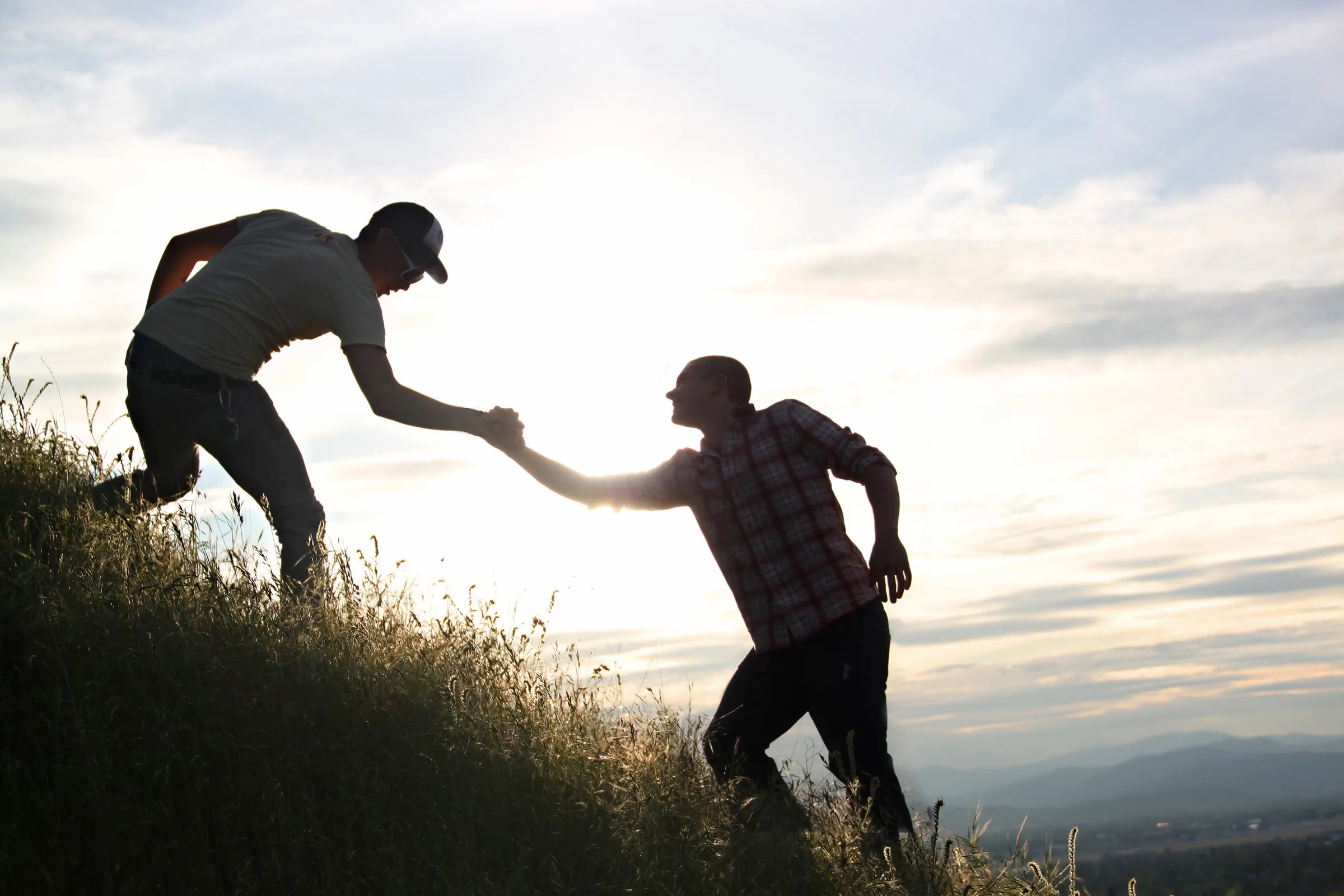 Stock photo of two men on a grassy hill at sunset, one reaching down to help the other up